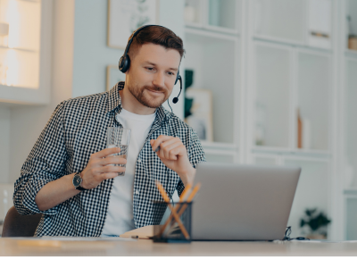 Ein lächelnder Mann im karierten Hemd mit Headset sitzt an einem Schreibtisch vor seinem Laptop. In der einen Hand hält er ein Glas Wasser, während er auf den Bildschirm blickt.