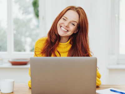 Junge Frau sitzt hinter einem Laptop und lächelt mit schräg gelegtem Kopf